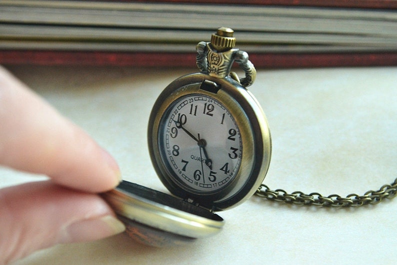 K&ouml;nnte beinhalten: Eine antike Taschenuhr aus Bronze mit rundem Geh&auml;use und passender Metallkette. Das wei&szlig;e Zifferblatt zeigt schwarze arabische Ziffern von 1 bis 12, einen inneren Ring mit Zahlen von 13 bis 24, schwarze Zeiger und das Wort QUARTZ.