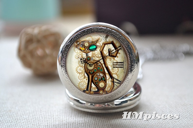 K&ouml;nnte beinhalten: Ein silberfarbener Taschenuhr-Anh&auml;nger mit einer Katzenillustration, die aus Zahnr&auml;dern und Uhrwerksteilen besteht. Die Katze hat ein gelbes und ein gr&uuml;nes Auge. Der Text HMpisces ist in der unteren rechten Ecke des Bildes aufgedruckt.
