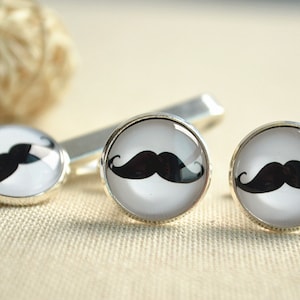 May include: A set of silver-toned cufflinks, a tie clip, and a pair of cufflinks, each featuring a black mustache design on a white background. The accessories are displayed on a neutral-toned surface.