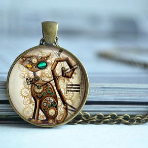 K&ouml;nnte beinhalten: Ein runder Anh&auml;nger in Bronzefarbe mit einer Katzenillustration aus mechanischen Zahnr&auml;dern, Zifferbl&auml;ttern und Federn. Die Katze hat ein gelbes und ein gr&uuml;nes Auge vor einem cremefarbenen Hintergrund mit r&ouml;mischen Ziffern.