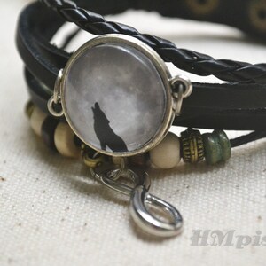 May include: A black leather bracelet with a silver charm featuring a wolf howling at the moon. The bracelet also has brown and green beads.