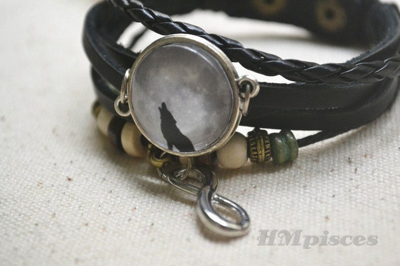 May include: A black leather bracelet with a silver charm featuring a wolf howling at the moon. The bracelet also has brown and green beads.
