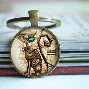 K&ouml;nnte beinhalten: Ein runder Schl&uuml;sselanh&auml;nger in Bronzefarbe mit einem Glaskabochon, der eine Katze im Steampunk-Stil zeigt. Die Katze besteht aus verschiedenen mechanischen Zahnr&auml;dern, Uhrteilen und r&ouml;mischen Ziffern in Braun-, Gold- und T&uuml;rkist&ouml;nen auf cremefarbenem Grund.