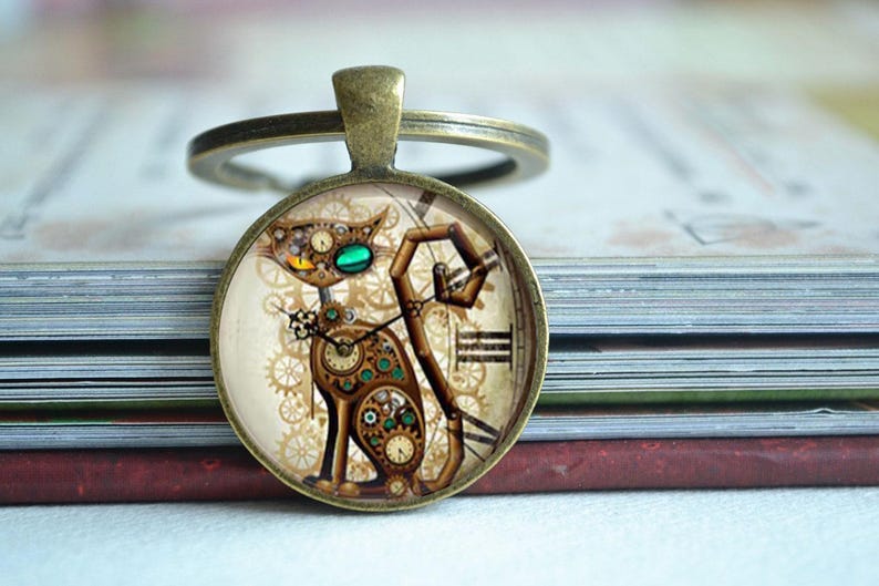 K&ouml;nnte beinhalten: Ein runder Schl&uuml;sselanh&auml;nger in Bronzefarbe mit einem Glaskabochon, der eine Katze im Steampunk-Stil zeigt. Die Katze besteht aus verschiedenen mechanischen Zahnr&auml;dern, Uhrteilen und r&ouml;mischen Ziffern in Braun-, Gold- und T&uuml;rkist&ouml;nen auf cremefarbenem Grund.