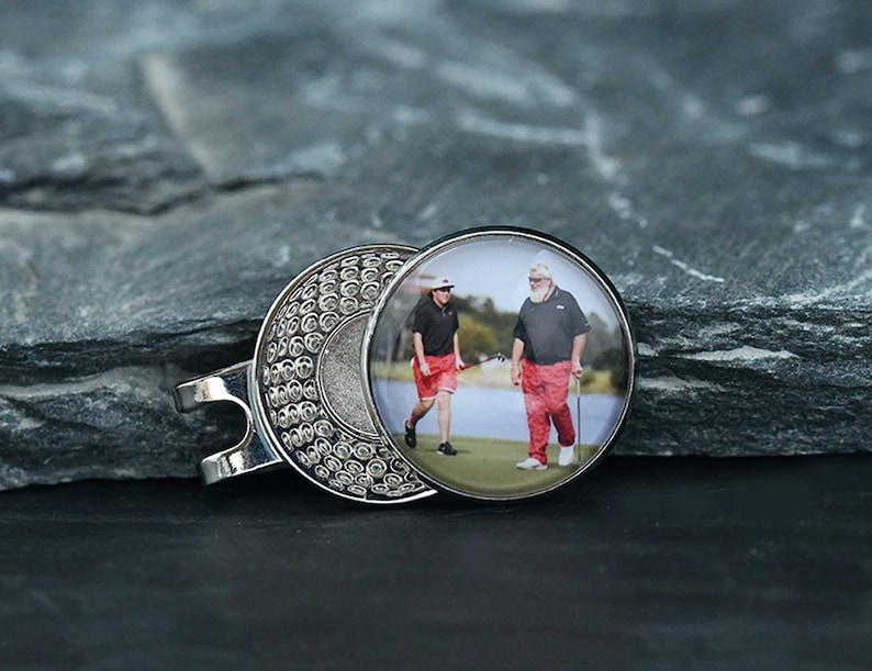 May include: Silver golf ball marker with a round photo insert featuring two people wearing red shirts and holding golf clubs. The photo is of two people on a golf course.