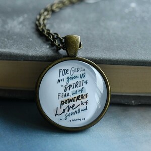May include: A round pendant necklace with a bronze-colored chain. The pendant has a white background with black handwritten text. The text reads "For God has not given us spirit of fear, but of power & love & of a sound mind."