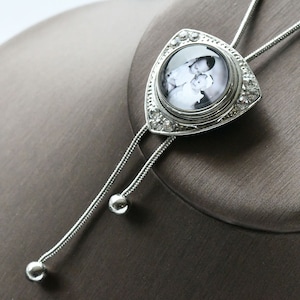 May include: A silver bolo tie with a triangular pendant featuring a black and white photo of two people. The pendant is decorated with clear rhinestones.