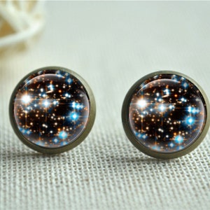 Puede incluir: Un par de pendientes de bot&oacute;n redondos con un dise&ntilde;o celestial. Los pendientes presentan un fondo oscuro con acentos blancos, naranjas y azules en forma de estrella, engastados en un metal color bronce.