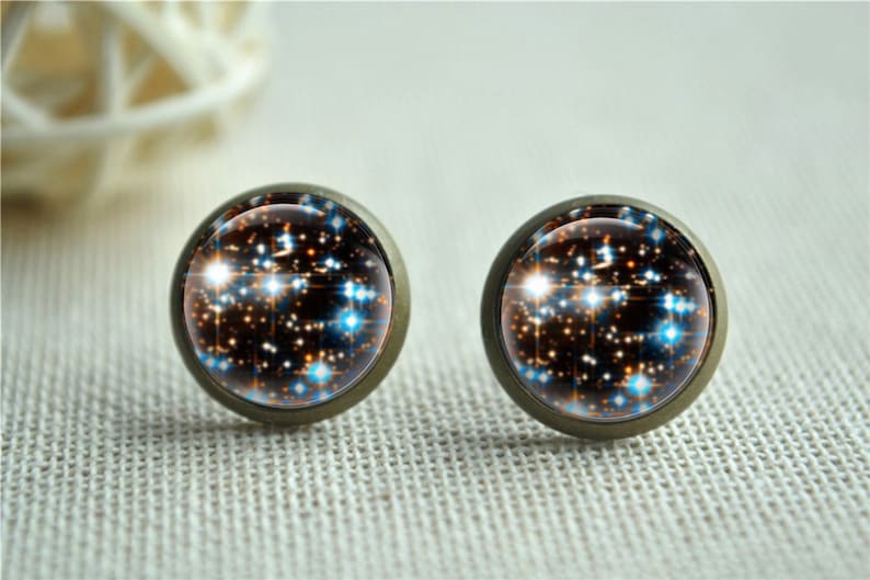 Puede incluir: Un par de pendientes de bot&oacute;n redondos con un dise&ntilde;o celestial. Los pendientes presentan un fondo oscuro con acentos blancos, naranjas y azules en forma de estrella, engastados en un metal color bronce.