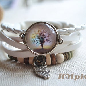 May include: White leather wrap bracelet with a round glass charm featuring a colorful tree image. The bracelet also has wooden beads and an owl charm.