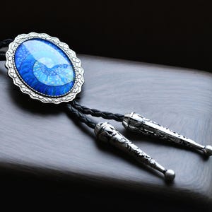 Puede incluir: Una corbata bolo con un colgante ovalado plateado con un diseño azul y blanco. El colgante tiene un borde plateado decorativo. Cordones trenzados negros conducen a puntas plateadas.