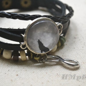 May include: A black leather bracelet with a silver charm featuring a wolf howling at the moon. The bracelet has a braided leather strap and silver beads.