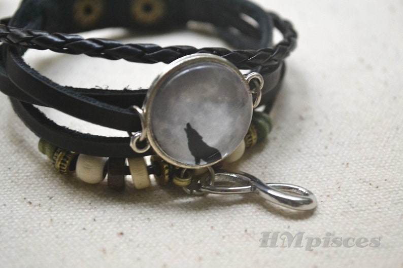 May include: A black leather bracelet with a silver charm featuring a wolf howling at the moon. The bracelet has a braided leather strap and silver beads.