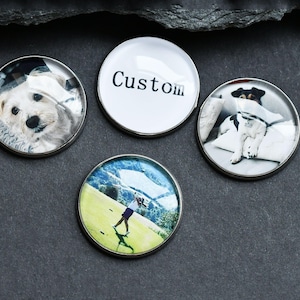 Pode incluir: Quatro marcadores de bola de golfe redondos de metal. Três marcadores apresentam imagens de um cão, um jogador de golfe e a palavra "Custom". Os marcadores têm um acabamento transparente e brilhante e estão sobre um fundo cinza escuro.