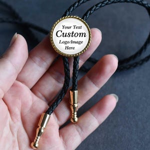 May include: A black bolo tie with a braided leather cord and gold-colored metal accents. The circular pendant has a gold-colored border and a white center with the text "Your Text Custom Logo/Image Here".
