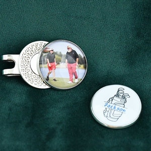 May include: A silver golf ball marker with a photo of two people on a golf course. The other side of the marker has a black and white illustration of a golf bag with the name "Jackson" written on it.