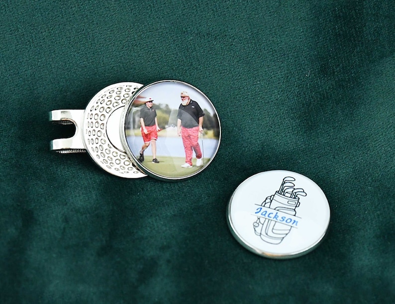 May include: A silver golf ball marker with a photo of two people on a golf course. The other side of the marker has a black and white illustration of a golf bag with the name "Jackson" written on it.