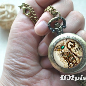 K&ouml;nnte beinhalten: Ein bronzefarbener Taschenuhr-Anh&auml;nger an einer passenden Kette mit einem runden Zifferblatt, das eine mechanische Katzenillustration, komplexe Zahnradmuster, ein leuchtend gr&uuml;nes Auge und den wei&szlig;en Schriftzug HMpisces am unteren Rand zeigt.