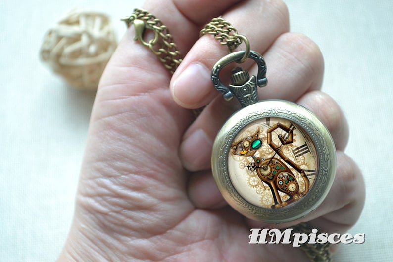K&ouml;nnte beinhalten: Ein bronzefarbener Taschenuhr-Anh&auml;nger an einer passenden Kette mit einem runden Zifferblatt, das eine mechanische Katzenillustration, komplexe Zahnradmuster, ein leuchtend gr&uuml;nes Auge und den wei&szlig;en Schriftzug HMpisces am unteren Rand zeigt.