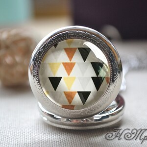Op de afbeelding: Een zilverkleurige ronde medaillonhanger met een glazen cabochon-inzetstuk. Het inzetstuk toont een herhalend geometrisch patroon van driehoeken in tinten zwart, mosterdgeel, roestoranje en wit op een lichtgrijze achtergrond.