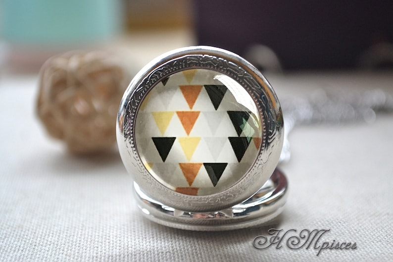 Op de afbeelding: Een zilverkleurige ronde medaillonhanger met een glazen cabochon-inzetstuk. Het inzetstuk toont een herhalend geometrisch patroon van driehoeken in tinten zwart, mosterdgeel, roestoranje en wit op een lichtgrijze achtergrond.