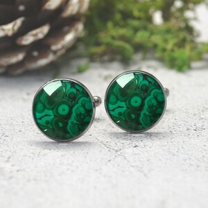 May include: A pair of round cufflinks featuring a polished green malachite stone inlay with distinct concentric circular patterns. The stones are set within a smooth, dark metallic frame, showcasing a vibrant emerald and forest green color palette.