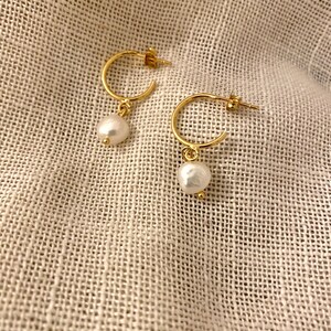 May include: A pair of gold hoop earrings with a single white pearl dangling from each hoop.