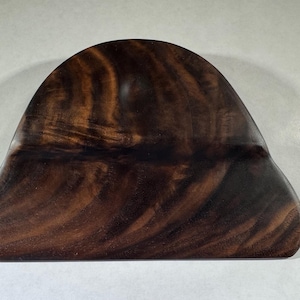 May include: A dark brown, wooden, hand-carved object with a smooth, curved shape. The wood grain is visible, creating a natural pattern. The object has a rounded top and a flat base.