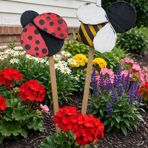 May include: Wooden garden stakes featuring a ladybug and a bee. The ladybug has a red body with black spots and black wings. The bee has a yellow and black striped body with white wings. The stakes are set in a flower bed.