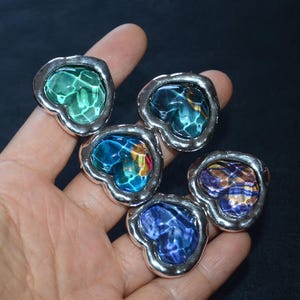 Könnte beinhalten: Fünf herzförmige Ringe mit silberfarbenen Rändern und farbenfrohen, strukturierten Glaszentren. Die Glasfarben umfassen Grün, Blau und Lila. Die Ringe werden auf dunklem Hintergrund präsentiert.