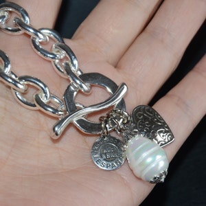 Puede incluir: Un brazalete de plata con cierre de palanca y tres dijes: un coraz&oacute;n, una perla y un dije redondo con el texto "Made in Spain".