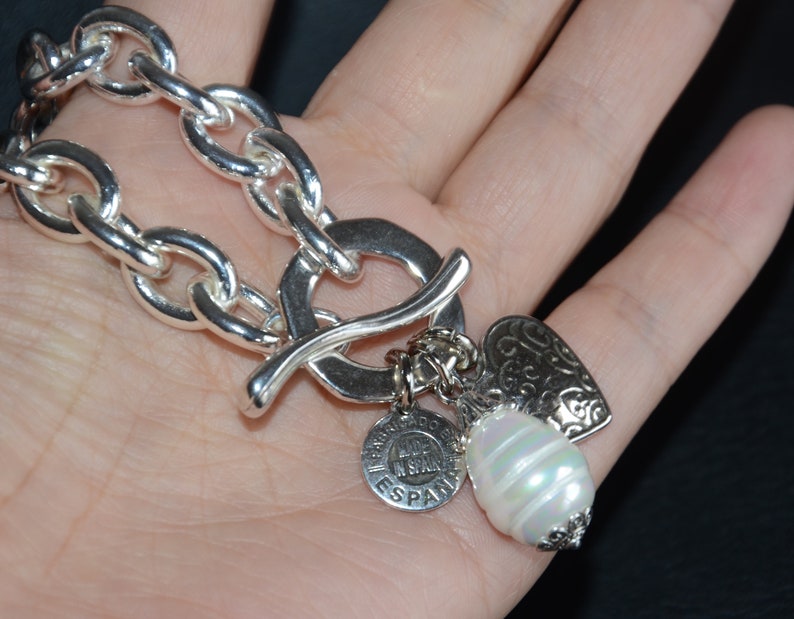 Puede incluir: Un brazalete de plata con cierre de palanca y tres dijes: un coraz&oacute;n, una perla y un dije redondo con el texto "Made in Spain".