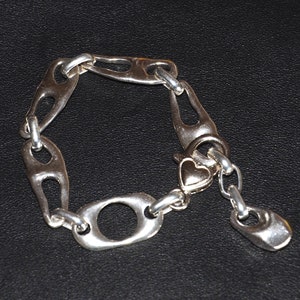 Puede incluir: Un brazalete de plata con un cierre en forma de corazón y un diseño de cadena de eslabones. El brazalete está hecho de eslabones de metal con forma de óvalos con recortes.