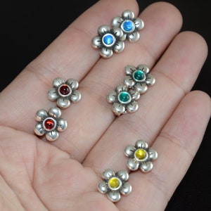 Puede incluir: Seis pares de pendientes de plata en forma de flor con piedras preciosas de diferentes colores. Los pendientes presentan un pequeño diseño de flor con una sola piedra preciosa en el centro. Las piedras preciosas son rojas, azules, verdes y amarillas.