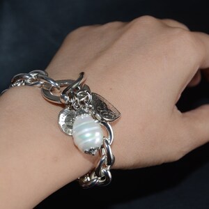 Puede incluir: Un brazalete de plata con un gran encanto de perla blanca y otros encantos m&aacute;s peque&ntilde;os. El brazalete est&aacute; en la mu&ntilde;eca de una persona.