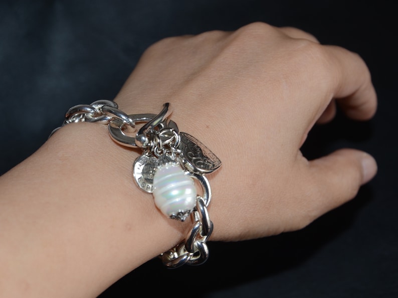 Puede incluir: Un brazalete de plata con un gran encanto de perla blanca y otros encantos m&aacute;s peque&ntilde;os. El brazalete est&aacute; en la mu&ntilde;eca de una persona.