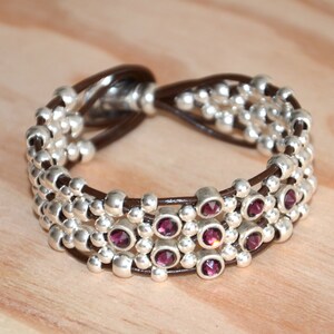 May include: A brown leather bracelet with silver beads and purple gemstones. The bracelet is made of multiple strands of leather and beads, with the gemstones interspersed throughout.