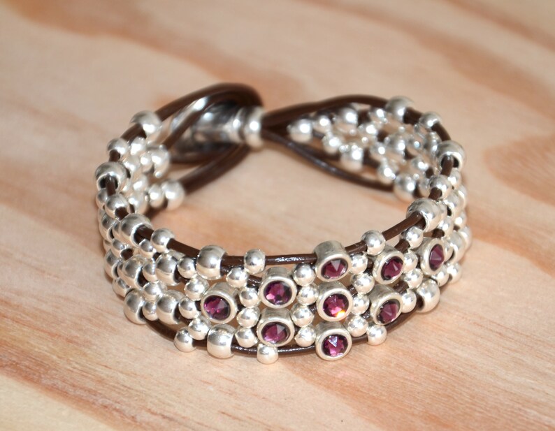 May include: A brown leather bracelet with silver beads and purple gemstones. The bracelet is made of multiple strands of leather and beads, with the gemstones interspersed throughout.