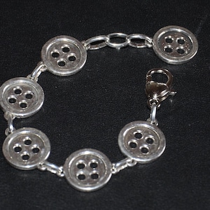 Könnte beinhalten: Ein silbernes Armband mit einer Kette und sechs Knopf-förmigen Anhängern. Jeder Anhänger hat vier Löcher in der Mitte.