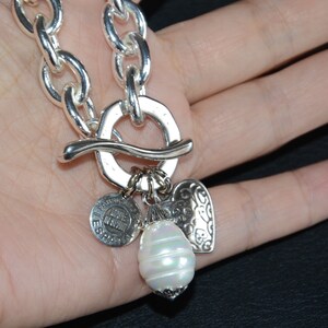 Puede incluir: Un collar de cadena de plata con un gran cierre de palanca de plata y varios colgantes. Los colgantes incluyen un colgante en forma de coraz&oacute;n, un colgante redondo con el texto "Made in Spain" y un gran colgante de perla blanca.