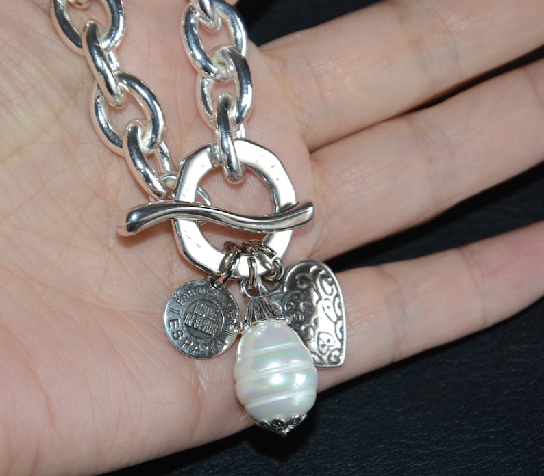 Puede incluir: Un collar de cadena de plata con un gran cierre de palanca de plata y varios colgantes. Los colgantes incluyen un colgante en forma de coraz&oacute;n, un colgante redondo con el texto "Made in Spain" y un gran colgante de perla blanca.