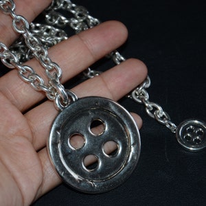 May include: A silver necklace featuring a large button pendant and a smaller button charm. The pendant has four holes in the centre and a textured edge.