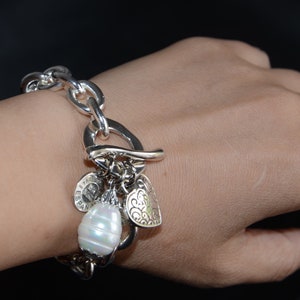 Puede incluir: Un brazalete de plata con una cadena de eslabones grandes de plata y varios abalorios, entre ellos un coraz&oacute;n, una perla y un abalorio redondo con la palabra "Espana" en &eacute;l.