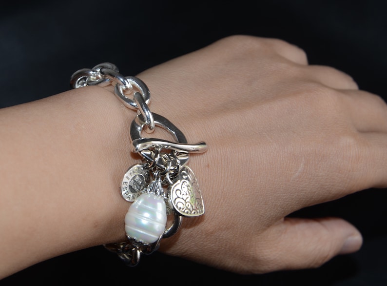 Puede incluir: Un brazalete de plata con una cadena de eslabones grandes de plata y varios abalorios, entre ellos un coraz&oacute;n, una perla y un abalorio redondo con la palabra "Espana" en &eacute;l.