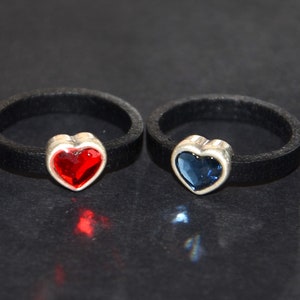 Może przedstawiać: Dwa czarne skórzane pierścienie z zawieszkami w kształcie serca. Jeden zawieszka ma czerwony kryształ, a drugi niebieski kryształ.