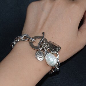 Puede incluir: Pulsera de plata con una cadena grande de plata, un dije de coraz&oacute;n, un dije redondo con la palabra "ESPANA" y un dije de perla blanca grande.