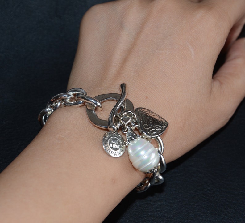 Puede incluir: Pulsera de plata con una cadena grande de plata, un dije de coraz&oacute;n, un dije redondo con la palabra "ESPANA" y un dije de perla blanca grande.