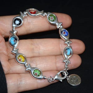 Può includere: Braccialetto d'argento con un ciondolo a forma di pesce con occhi di vetro colorati. Il braccialetto ha una chiusura a moschettone e un ciondolo con un design a forma di pesce.