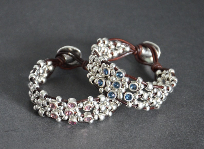 May include: Two leather bracelets with silver beads and colorful gemstones. The top bracelet has blue gemstones and the bottom bracelet has pink gemstones.