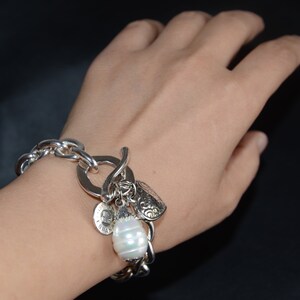 Puede incluir: Un brazalete de plata con un gran encanto de perla, un encanto en forma de coraz&oacute;n y otros encantos m&aacute;s peque&ntilde;os. El brazalete se lleva en la mu&ntilde;eca.